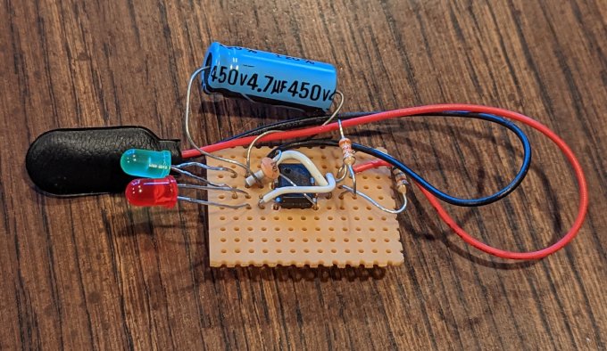 Hand-soldered NE555 circuit on perfboard, with red and green LEDs, resistors, capacitors, and a battery connector.